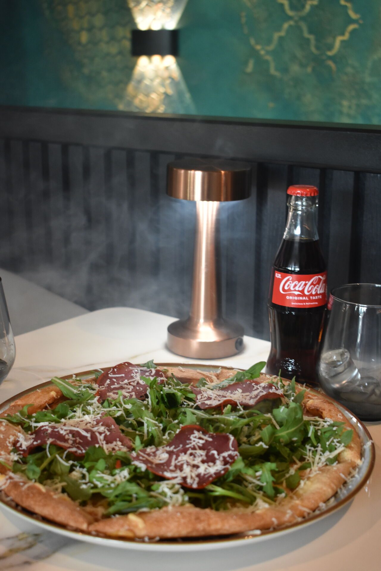 Pizza mit Rucola und Parmesan unter einer Kupferlampe, dahinter die Wand mit Goldornament